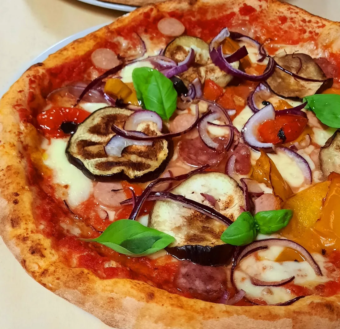 Pizza rossa con melanzane grigliate, peperoni, cipolla rossa, salame e basilico fresco.