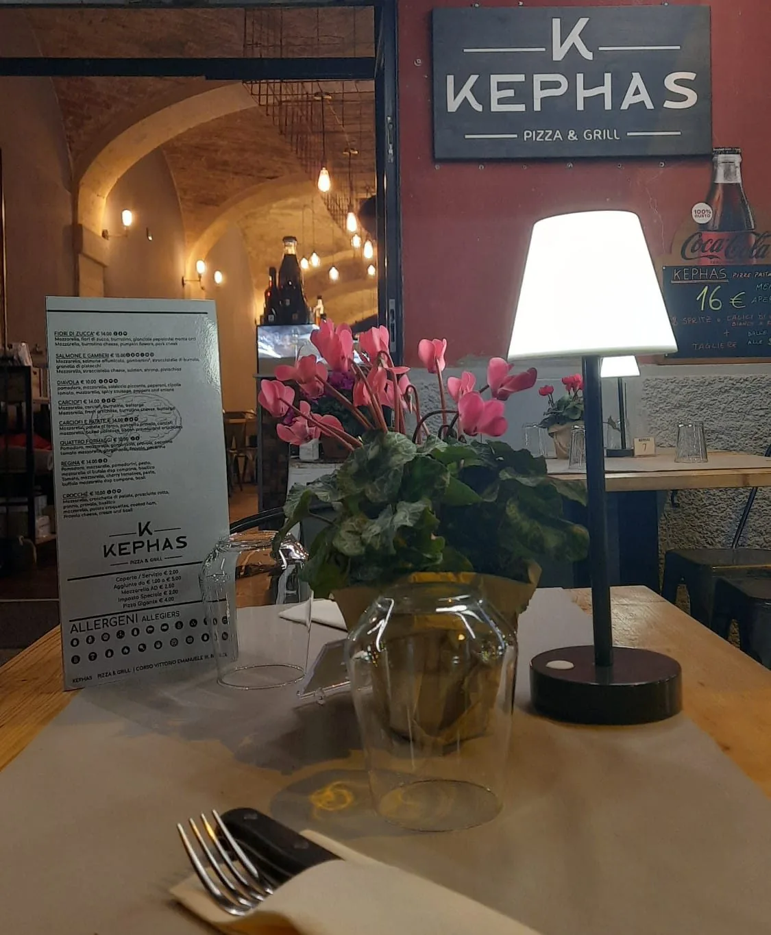 La sala di Kephas: insegna bianca su lastra scura appesa a un muro rosso mattone, e alle spalle la volta a botte in pietra illuminata da lampade appese.